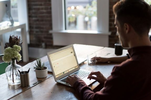 Person arbeitet am Laptop an einem Holztisch mit Pflanzen und einer Tasse Kaffee.